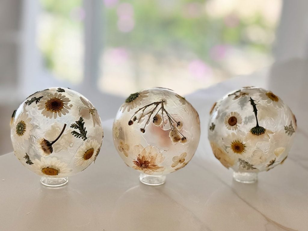 Three clear resin spheres containing dried flowers and leaves are displayed on a white surface with a blurred window and garden background.
