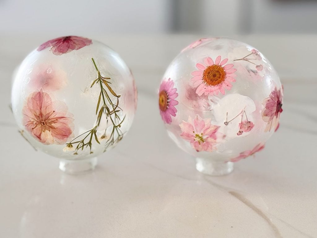 Two clear ice spheres with pink flowers and greenery frozen inside, resting on a smooth white surface.