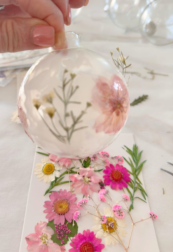 A hand holds a transparent ornament containing pressed flowers above a surface with more various pressed flowers and greenery.