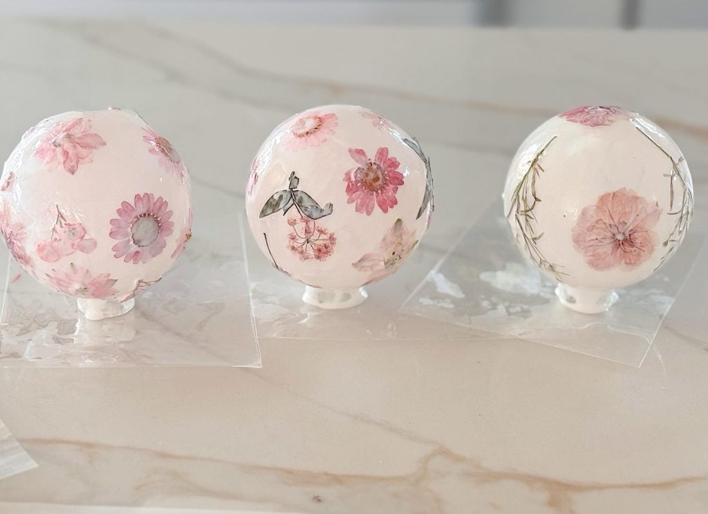 Three round objects with pink and purple floral designs are displayed on a marble surface, each resting on a clear square base.