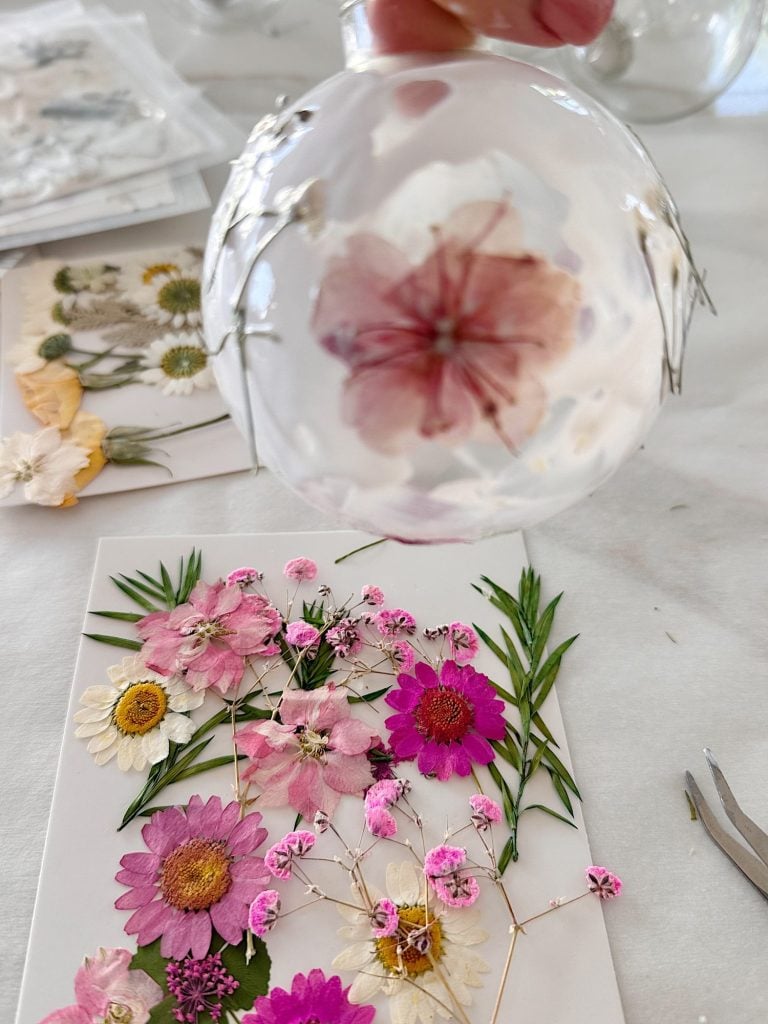 A hand holds a clear ornament with a pressed pink flower inside, above a table with various pressed flowers and crafting tools.