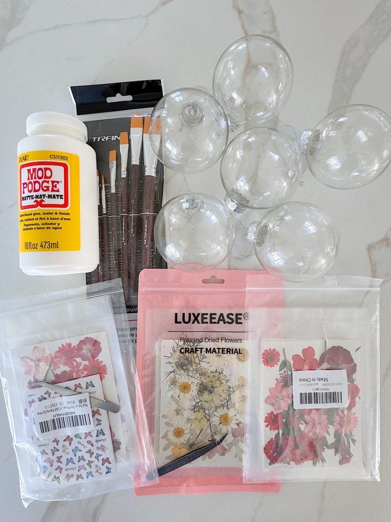 Clear plastic ornaments, Mod Podge, paint brushes, dried flowers, and botanical stickers arranged on a white surface for a craft project.
