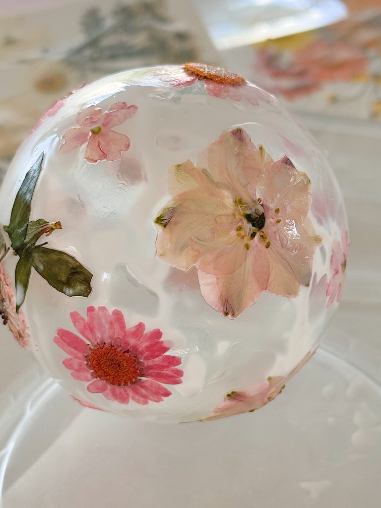 Clear sphere embedded with various dried flowers and petals, including pink and red blooms, resting on a transparent surface.