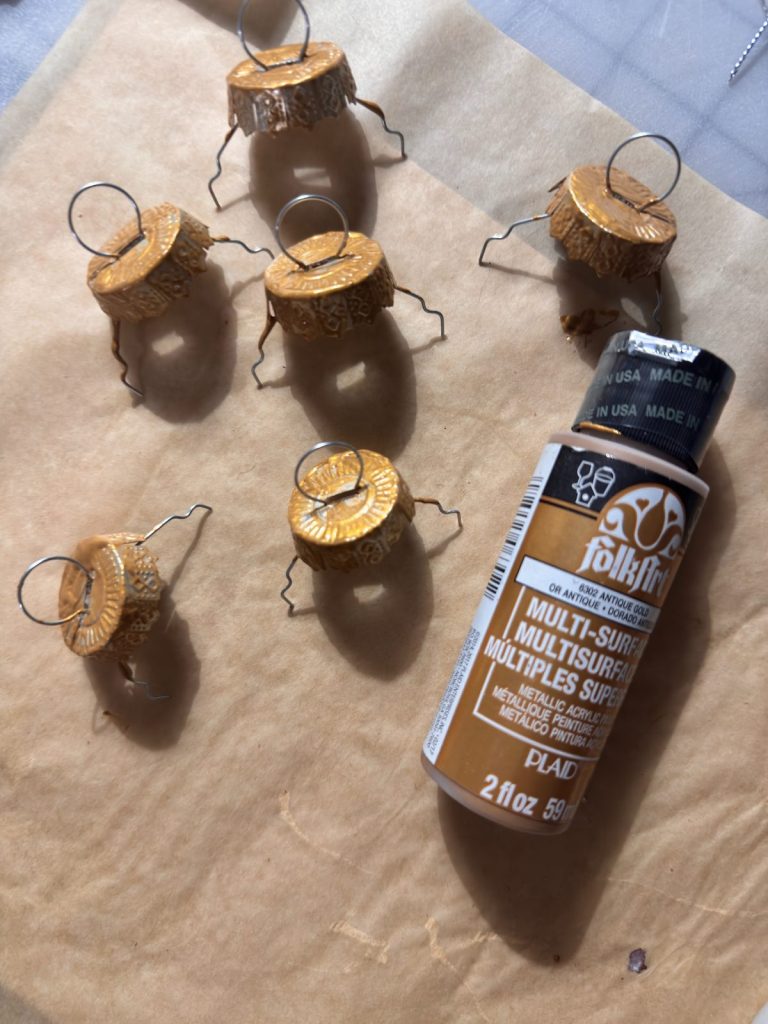 Six gold-painted bottle caps with wire stands—ideal for crafting glass ornaments—are drying on parchment paper next to a bottle of gold FolkArt multi-surface acrylic paint.