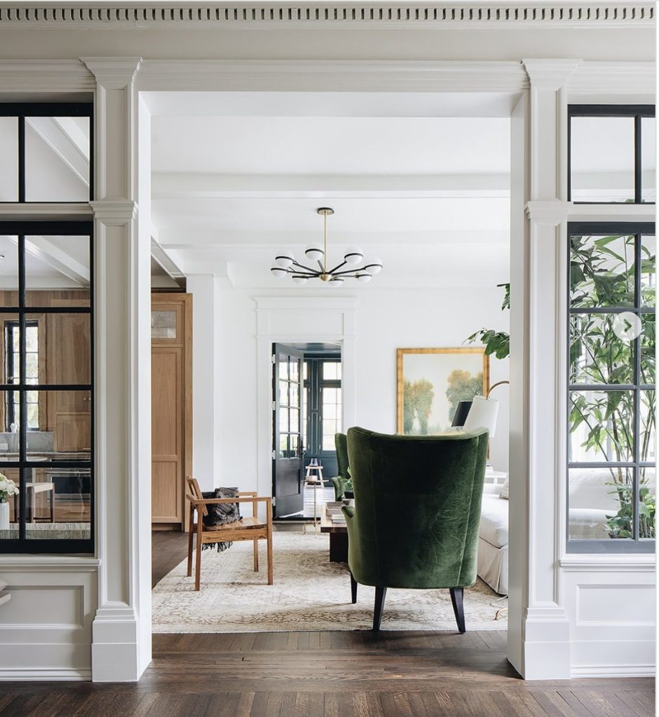 A bright living room with white walls, dark wood floors, a green velvet armchair, modern chandelier, and large windows separated by white trim.