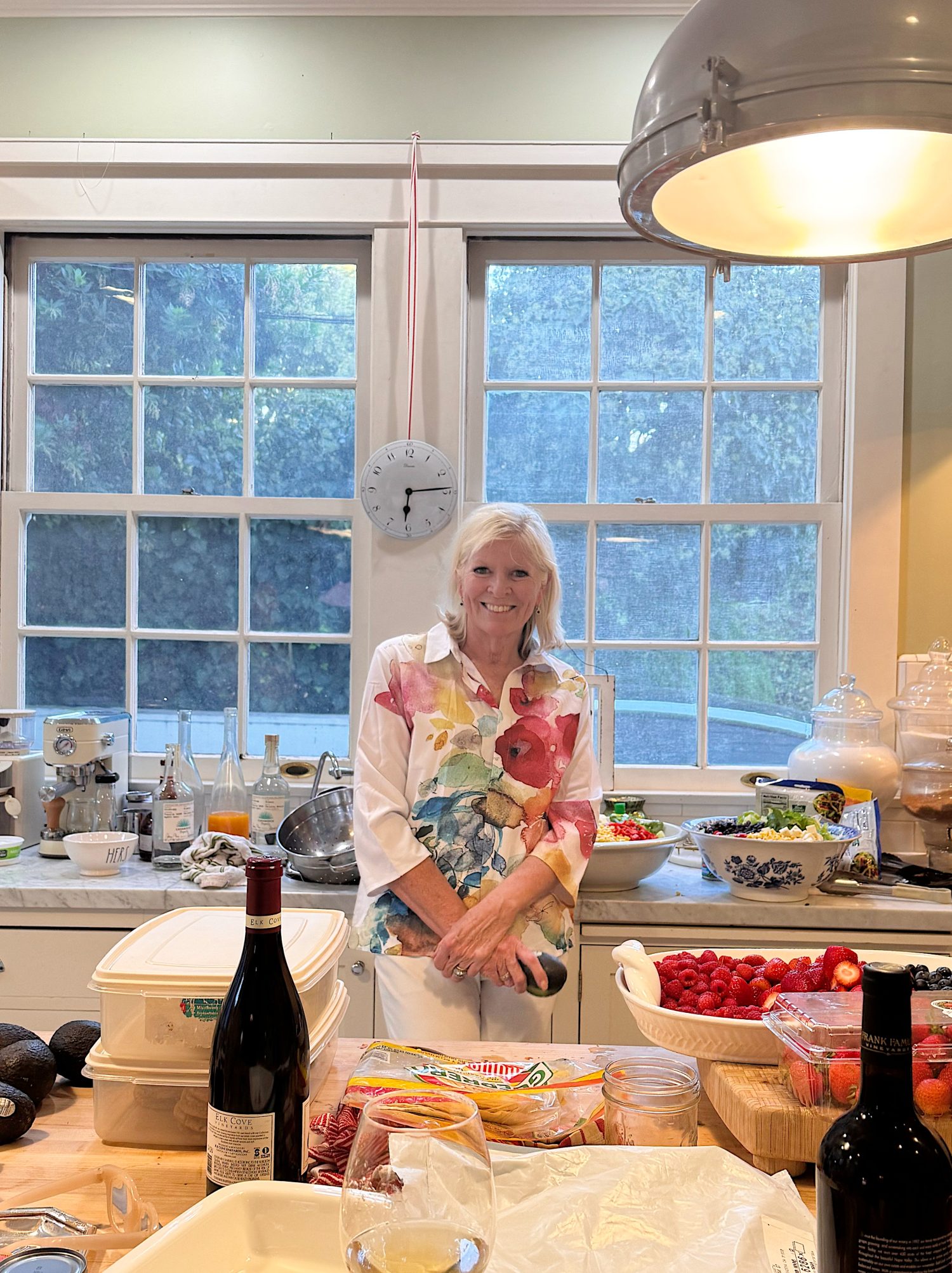 A woman stands in a bright kitchen filled with food, wine, and ingredients on the counter, with large windows and a clock in the background.