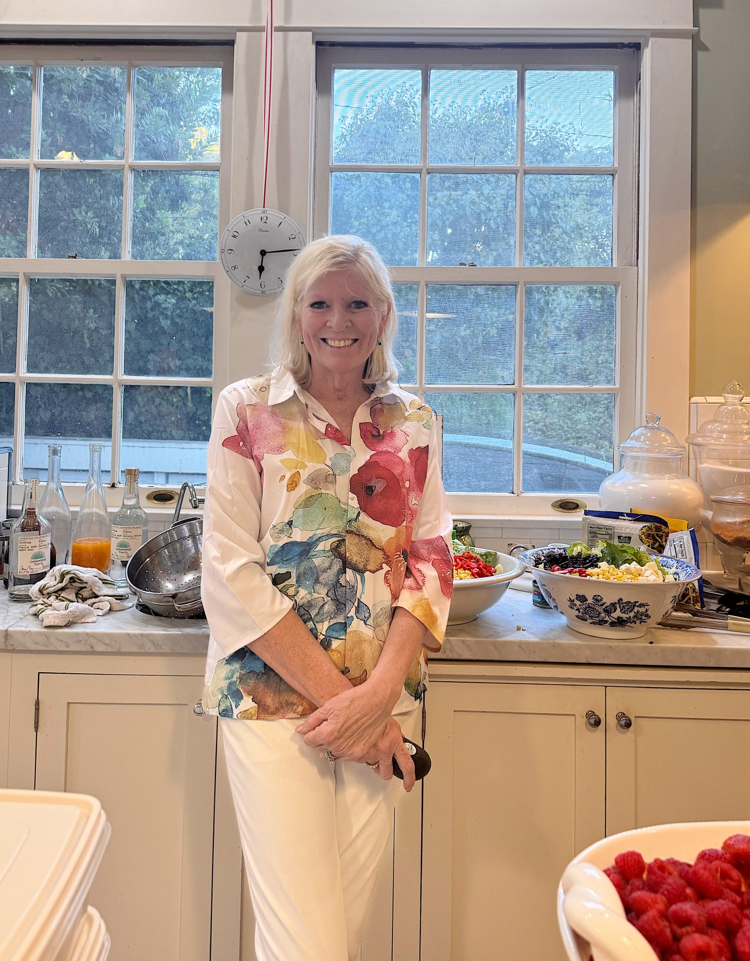 A woman with white hair stands in a kitchen in front of a counter with salad bowls, drinks, and food, wearing a floral shirt and white pants, smiling at the camera.