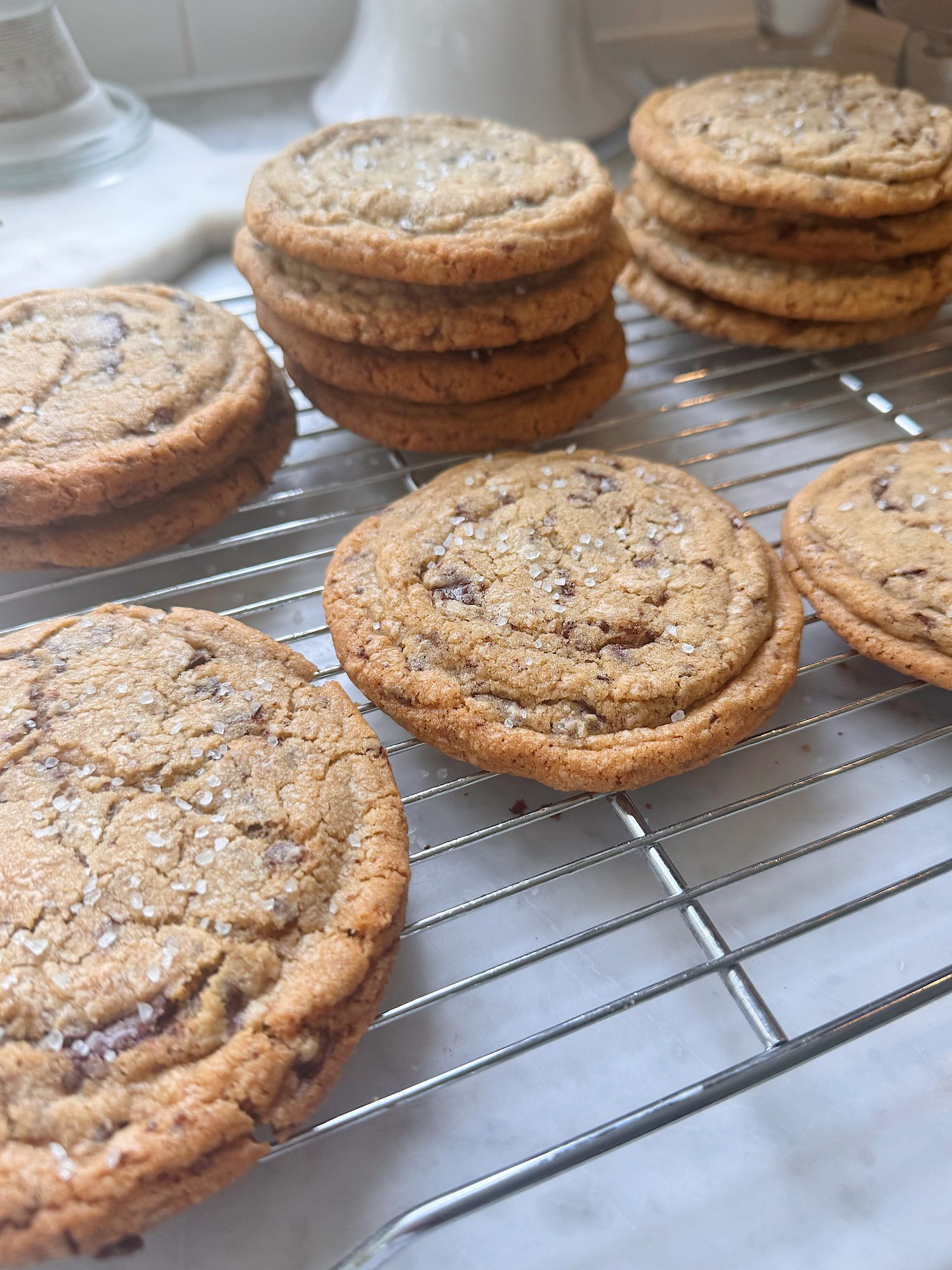 Stacks of chocolate chip cookies sprinkled with sea salt are cooling on a metal wire rack on a kitchen counter.