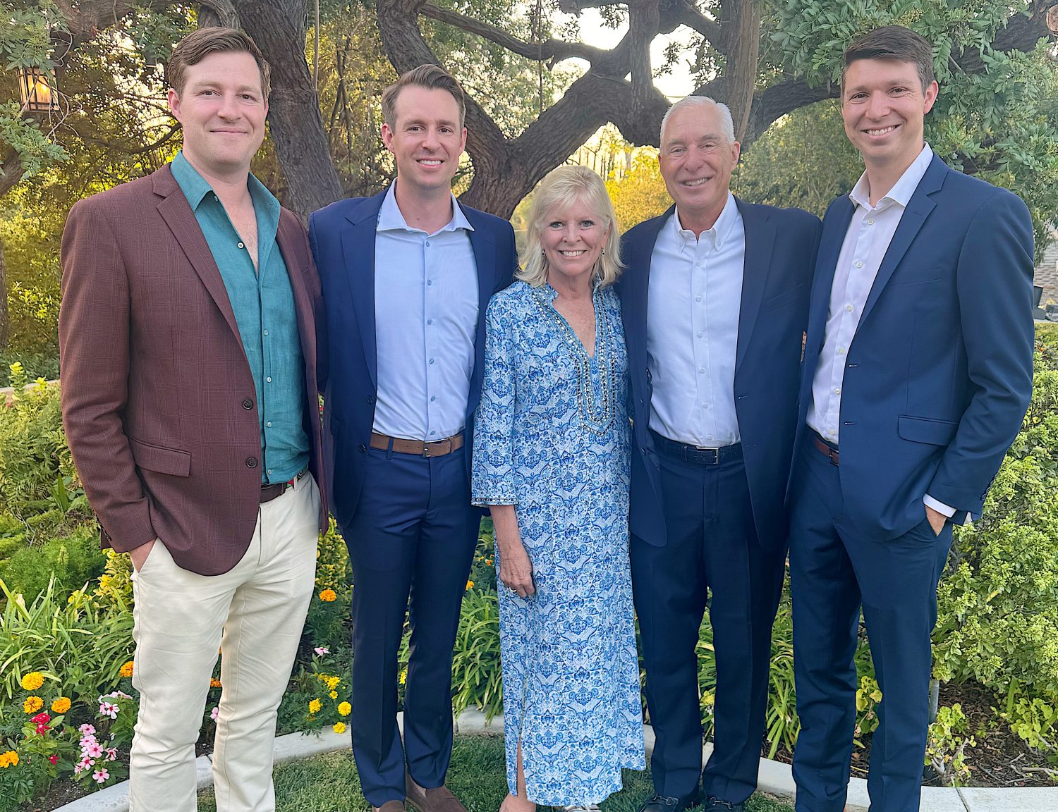 Five adults stand together outdoors in front of greenery and flowers, dressed in business and semi-formal attire, smiling at the camera.