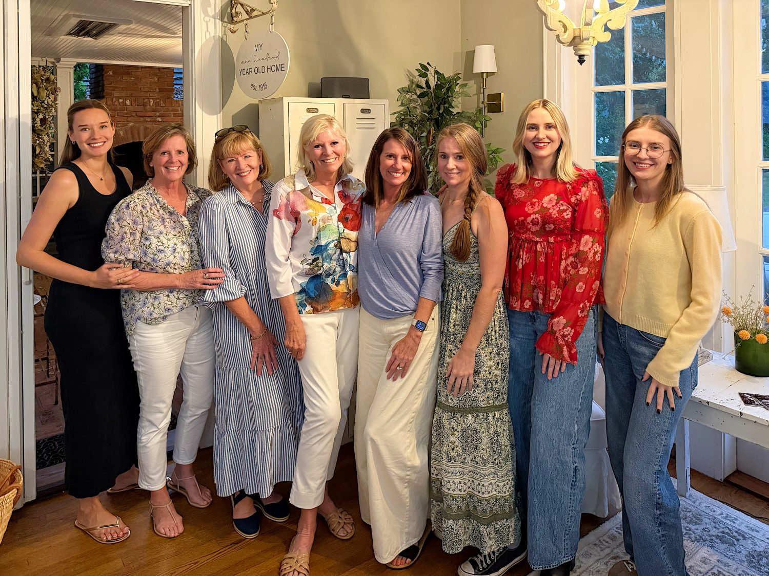 Eight women stand together indoors, smiling at the camera. They are dressed in casual, colorful clothing. The setting appears to be a cozy, well-lit living space.