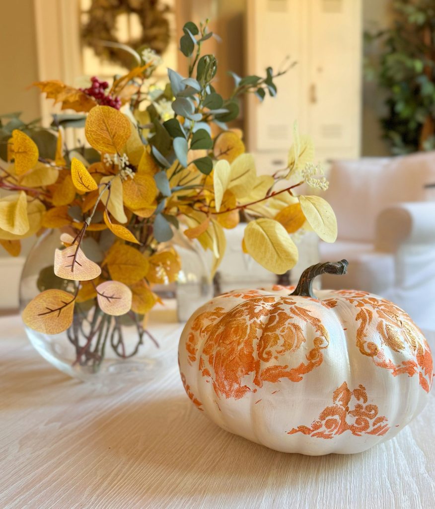 A white pumpkin with orange decorative patterns sits on a table next to a glass vase filled with autumn leaves.
