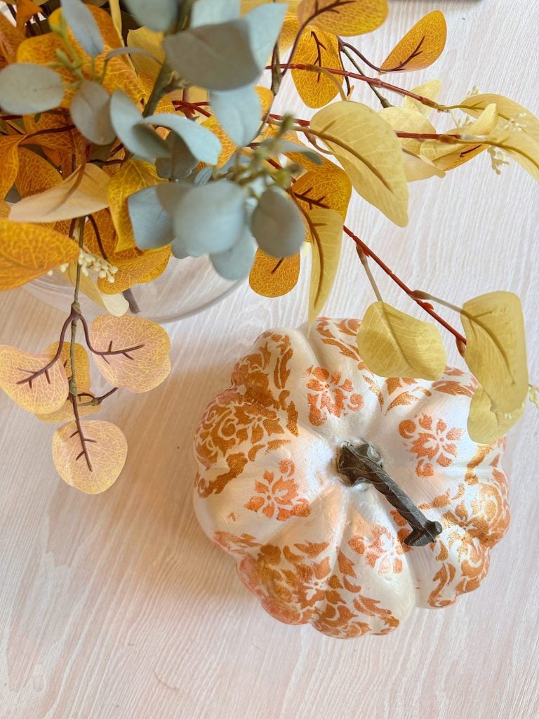 A white pumpkin with an orange floral pattern sits on a light wood surface beside a vase of yellow and gray artificial leaves.