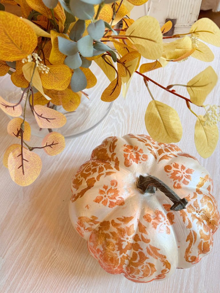 A white pumpkin with orange decorative patterns sits on a light wood surface next to a glass vase of yellow and green foliage.