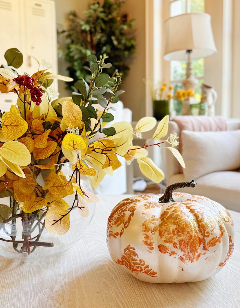 A white pumpkin with orange decorative patterns sits on a wooden table next to a glass vase of yellow autumn leaves in a bright, cozy living room.