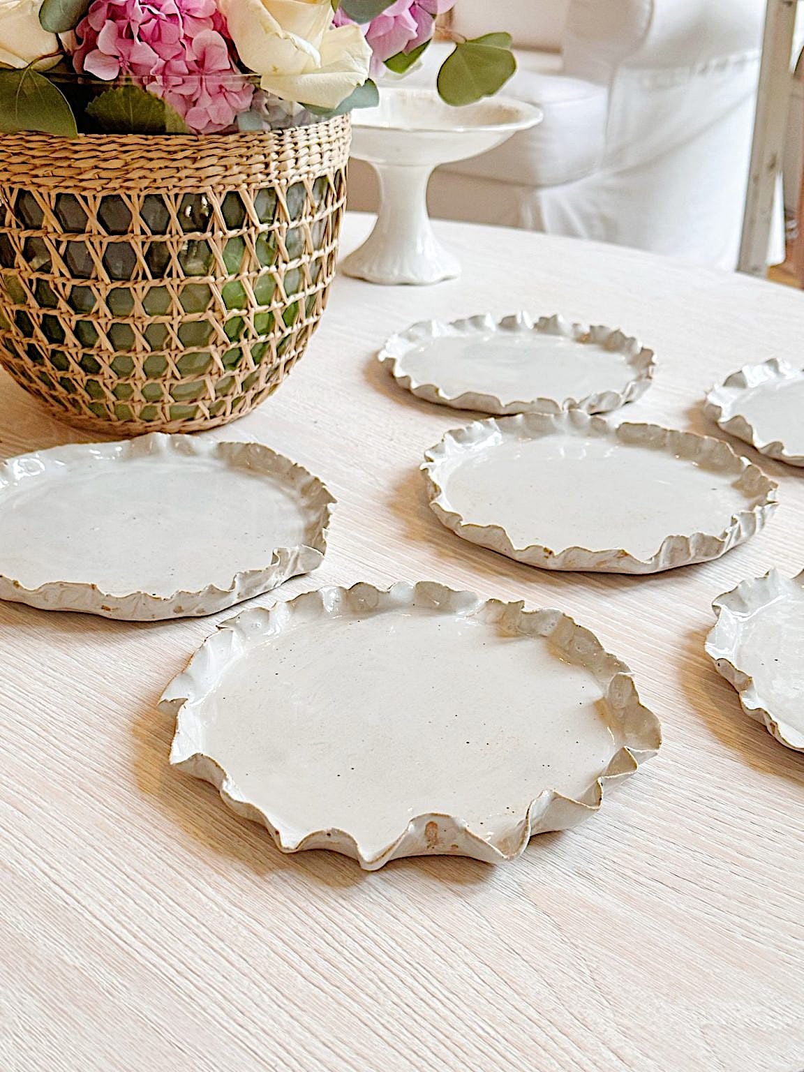 Six irregularly shaped, white ceramic plates are arranged on a light wooden table next to a woven basket with flowers.