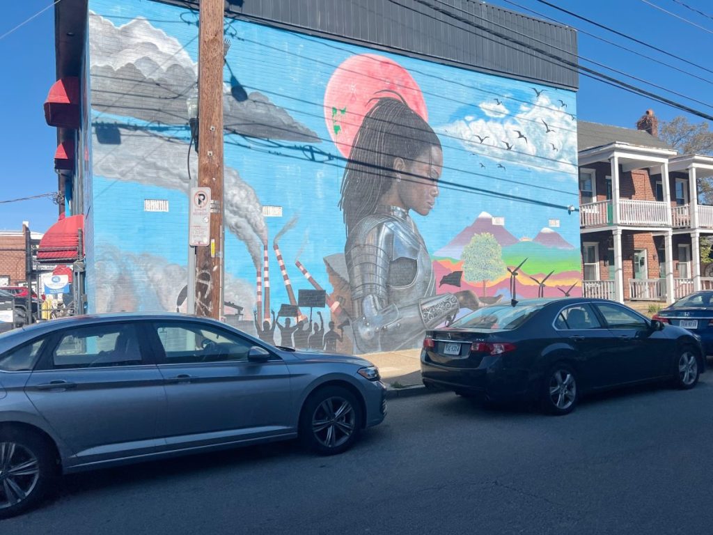 A mural of a woman in armor with braided hair is painted on the side of a building, with red sun and landscape in the background. Cars are parked along the street in front.