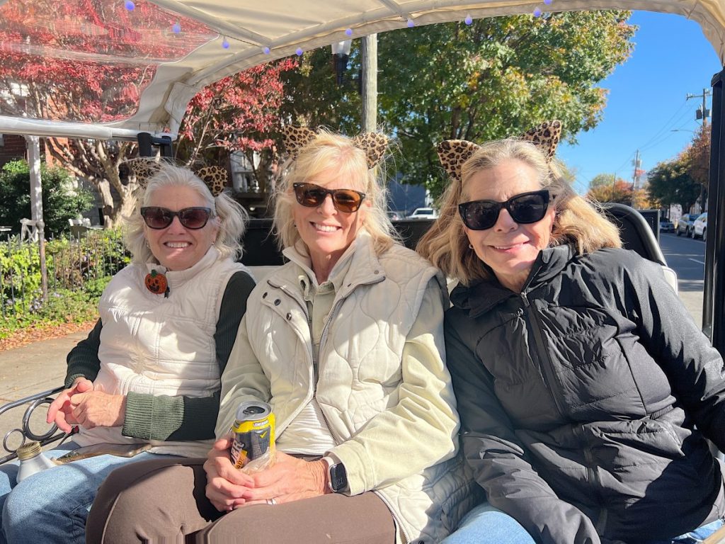 Three women wearing sunglasses and animal ear headbands sit together outdoors on a sunny day, smiling at the camera. One holds a can of drink. Autumn foliage is visible in the background.