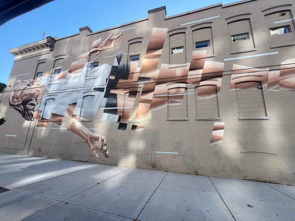 Large mural on a brick building depicts a fragmented, reclining human figure painted in a realistic style, with sections missing and aligned with window placements.