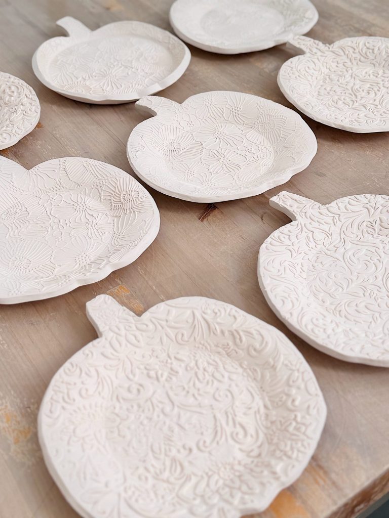 Several white ceramic plates shaped like pumpkins with detailed embossed floral patterns are arranged on a wooden surface.