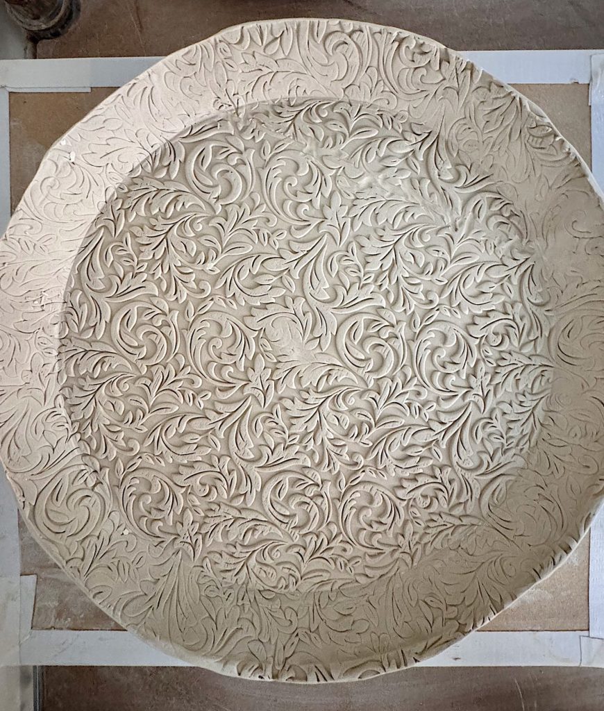A large, round clay plate with intricate, raised floral and leaf patterns covering the entire surface.