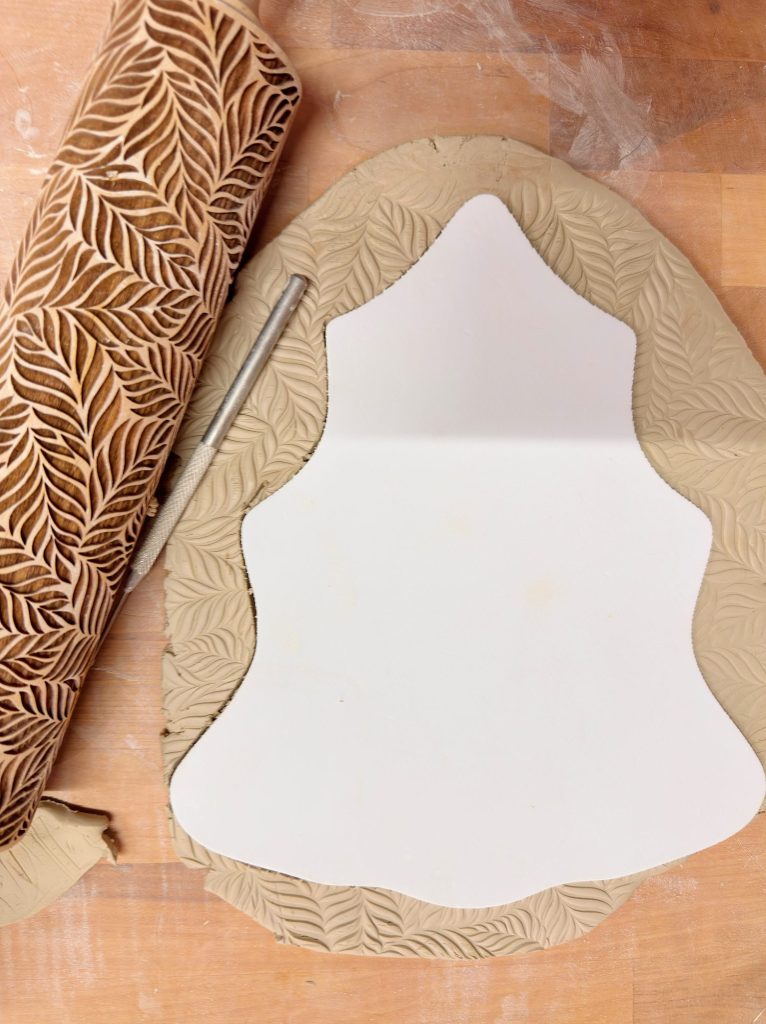 A sheet of clay with a leaf pattern is rolled out on a wooden surface, with a white template and a cutting tool placed on top.