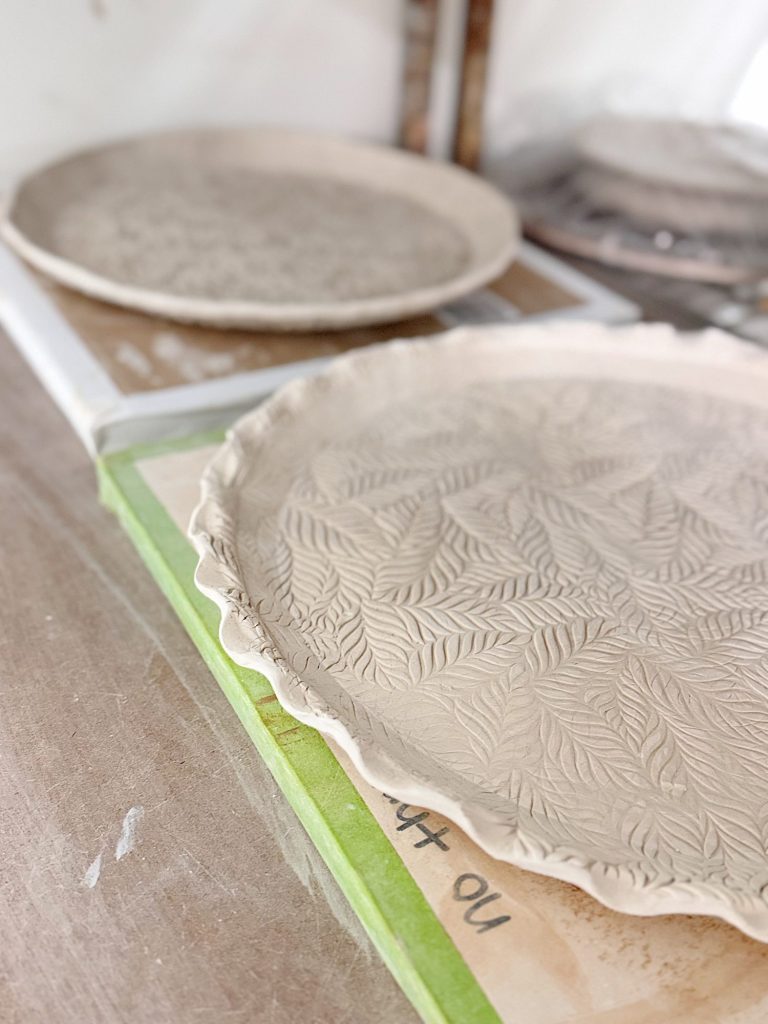 A close-up of two round, unglazed clay plates with leaf patterns pressed into the surface, resting on a work table in a pottery studio.