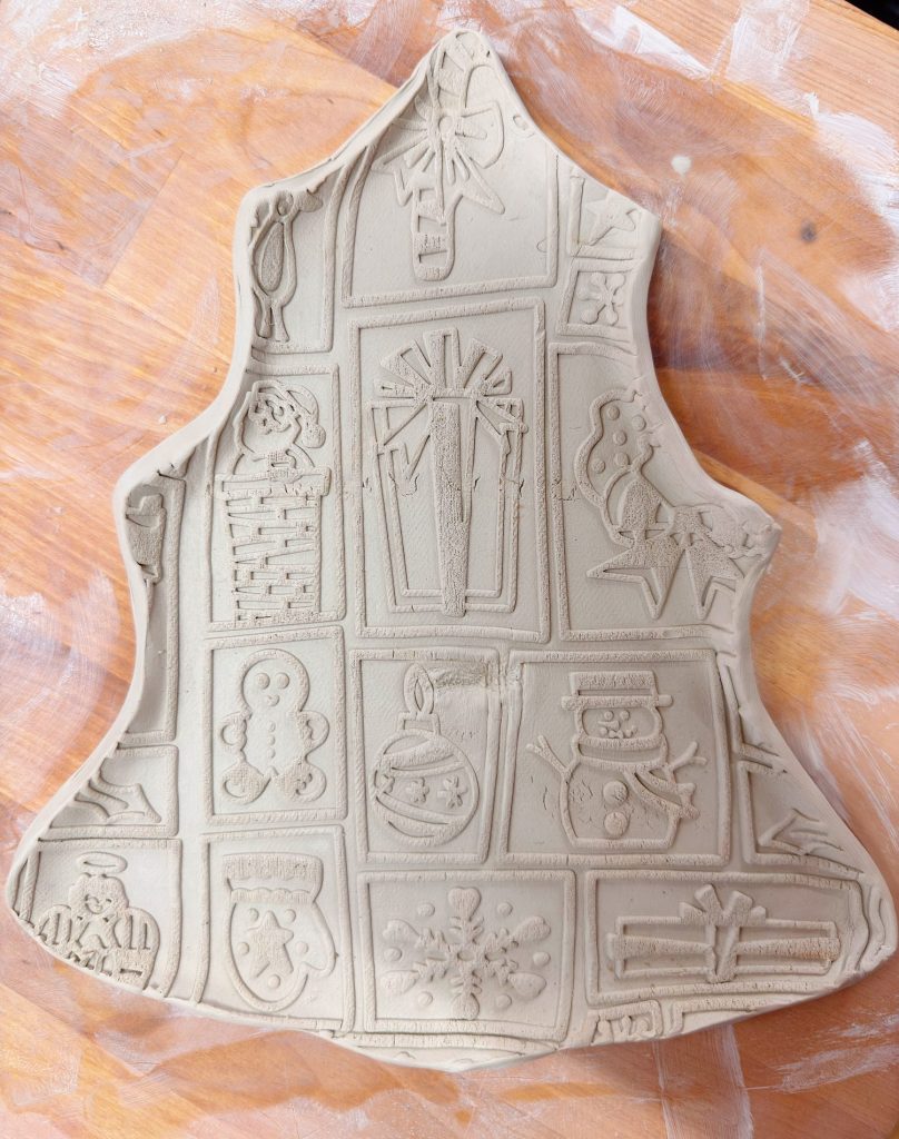 Piece of clay shaped like a Christmas tree, pressed with festive designs including presents, a snowman, gingerbread, ornaments, and a stocking, on a wooden surface dusted with flour or clay.