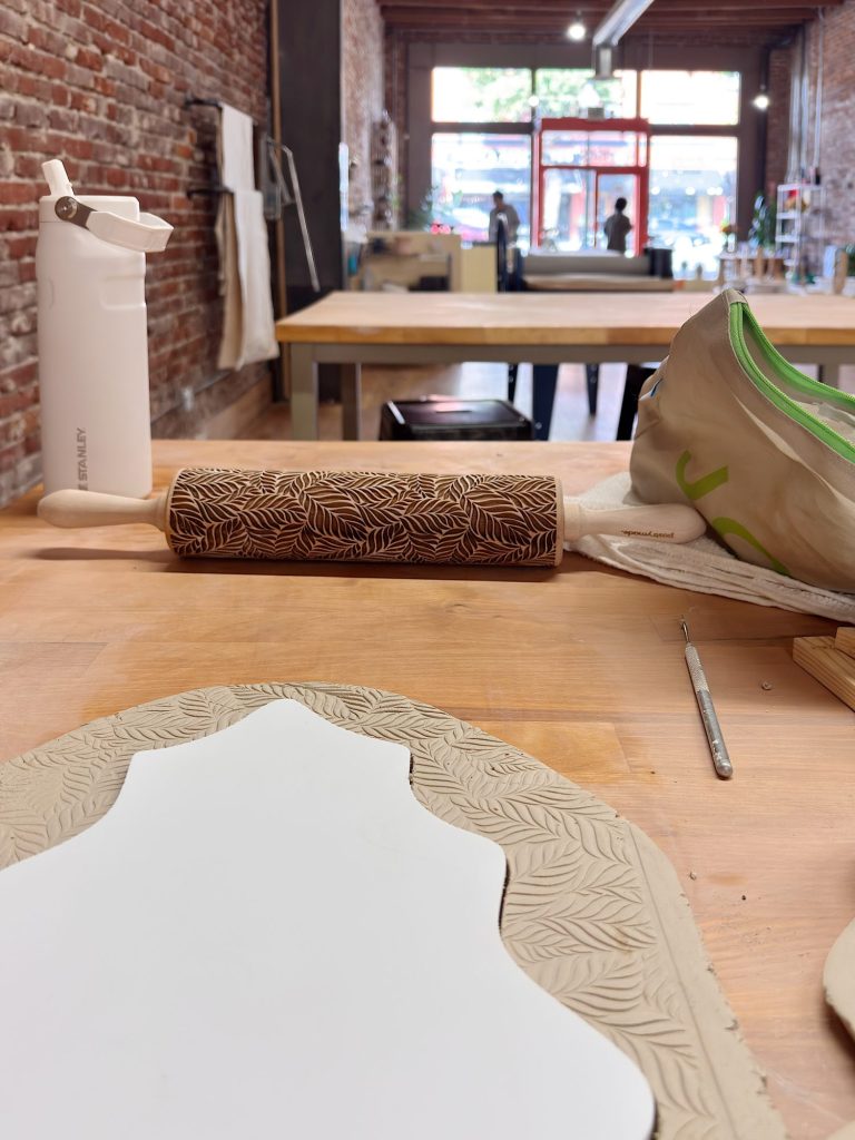 A rolling pin with a leaf pattern, a clay slab with the same pattern, a water bottle, and a tool sit on a wooden table in a studio with brick walls.