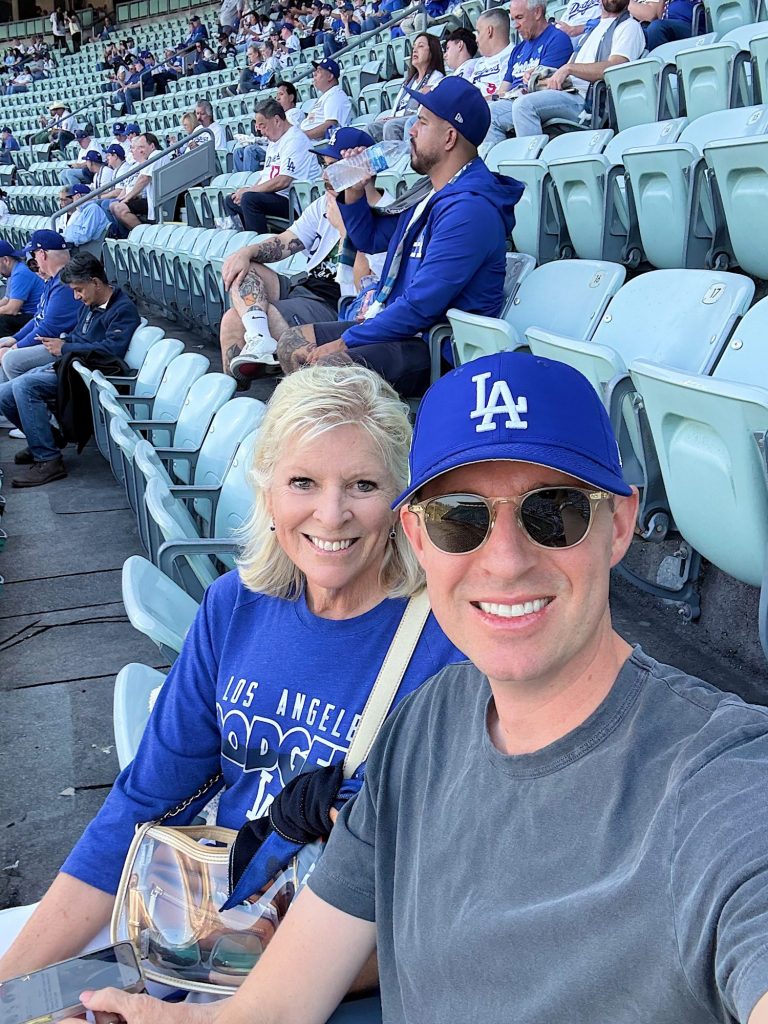 Two people sit in a stadium wearing Los Angeles Dodgers gear, smiling at the camera. Other fans are seated behind them in the stands.