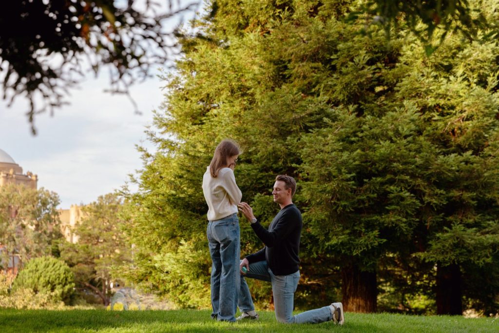 A man kneels on one knee, holding a woman's hand in a grassy park with tall trees in the background.