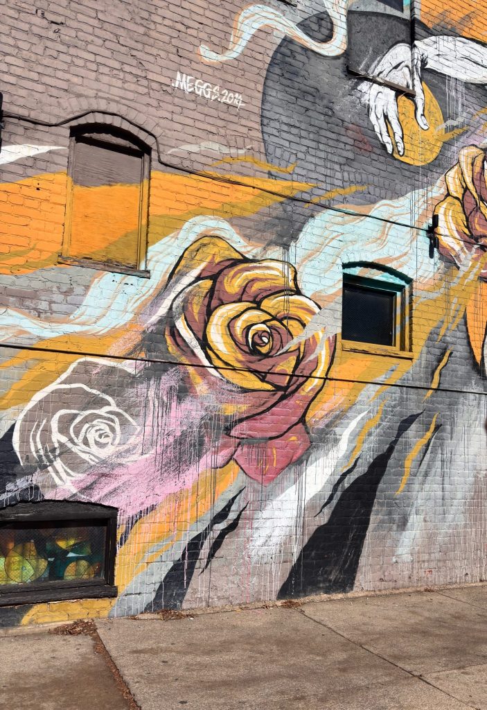 A mural painted on a brick wall features large, colorful roses and abstract shapes with black-trimmed windows integrated into the artwork.