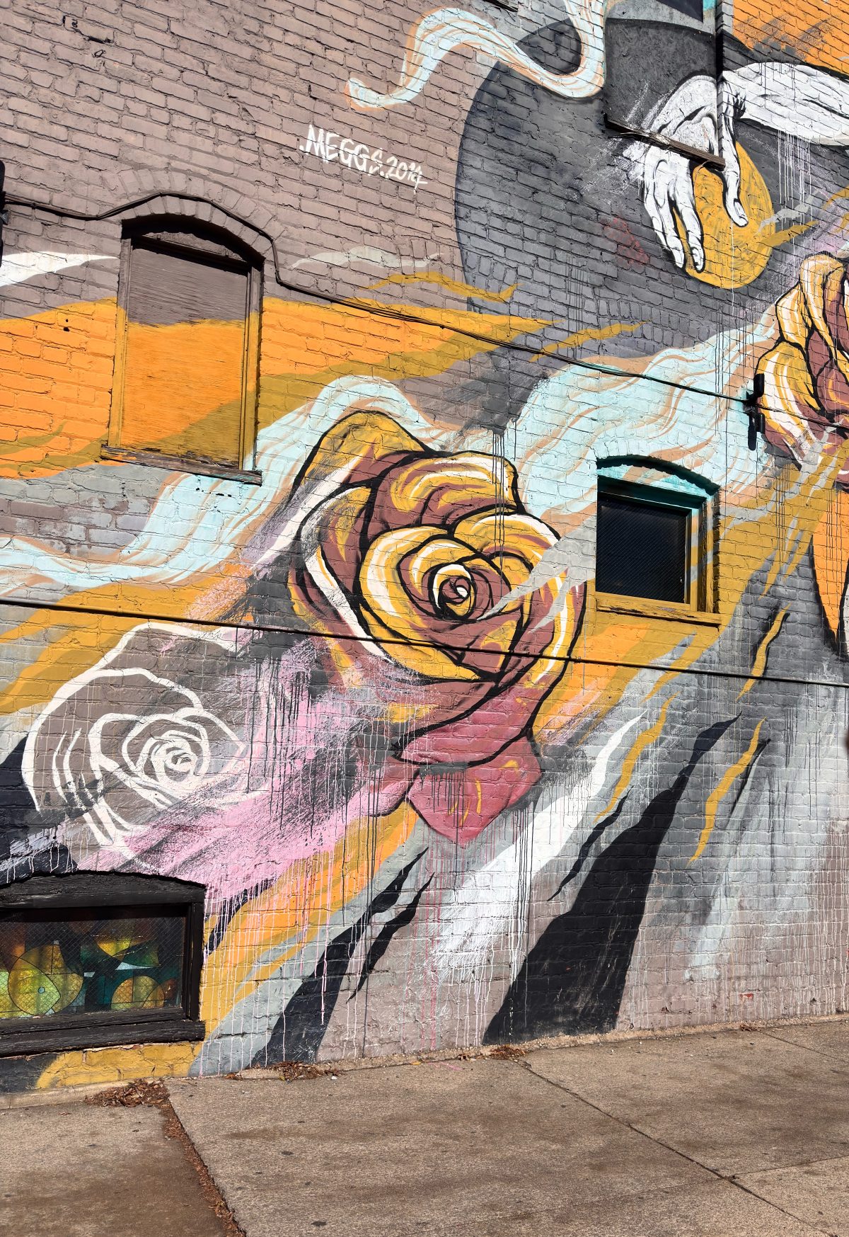 A mural painted on a brick wall features large, colorful roses and abstract shapes with black-trimmed windows integrated into the artwork.