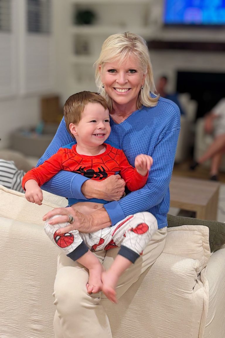 A smiling woman in a blue sweater sits on a couch holding a young child in red Spiderman pajamas, capturing a warm moment indoors during the holiday season. The background is softly blurred.