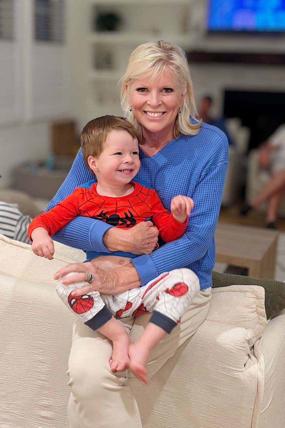 A smiling woman in a blue sweater sits on a couch and holds a young child wearing red Spiderman pajamas.