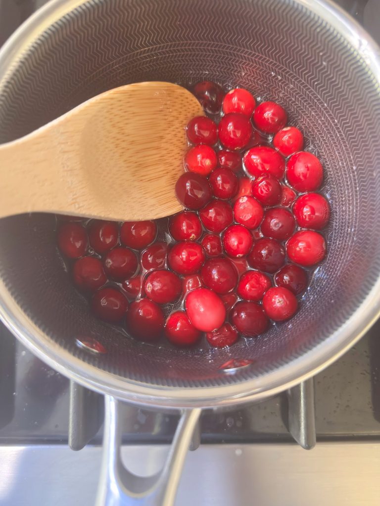 A wooden spoon stirring whole cranberries in a saucepan on a stove.