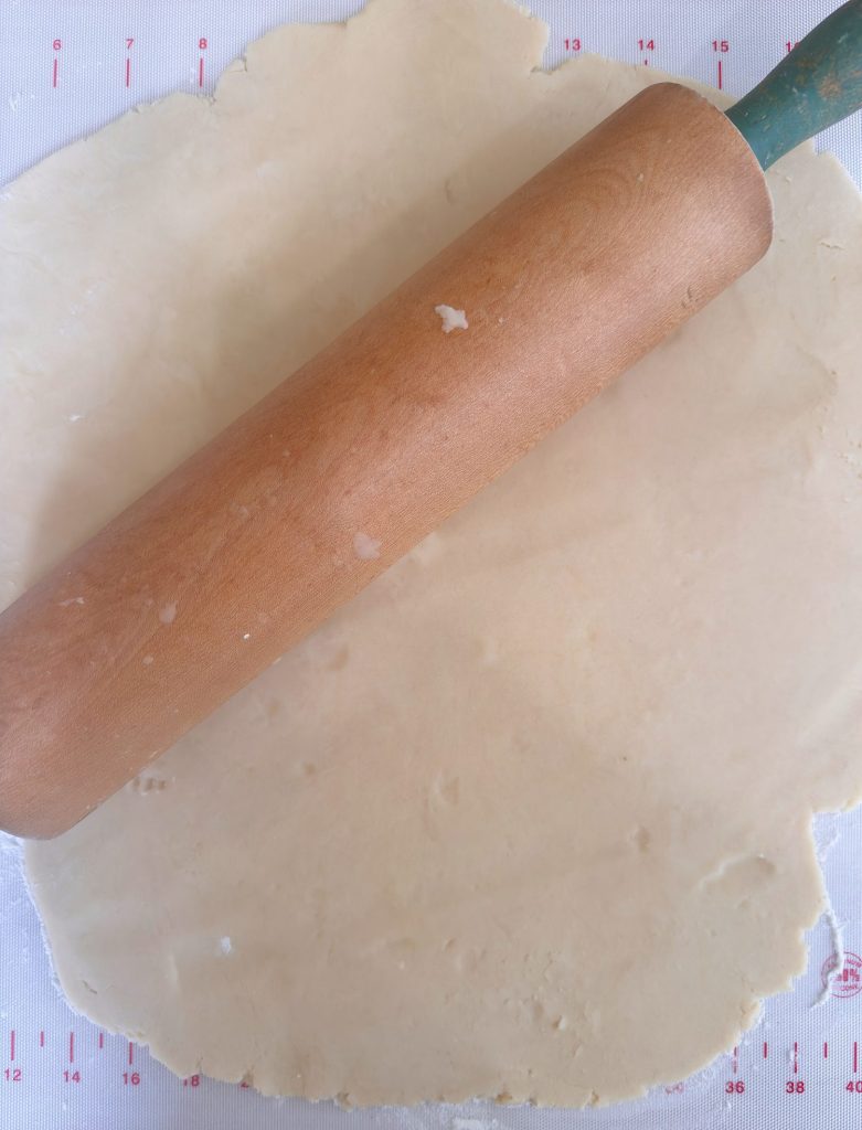 A wooden rolling pin is shown rolling out a sheet of dough on a lightly floured surface with measurement markings.