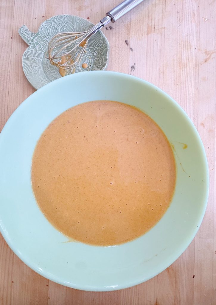 A whisk rests on a small plate beside a mixing bowl filled with light brown batter on a wooden surface.