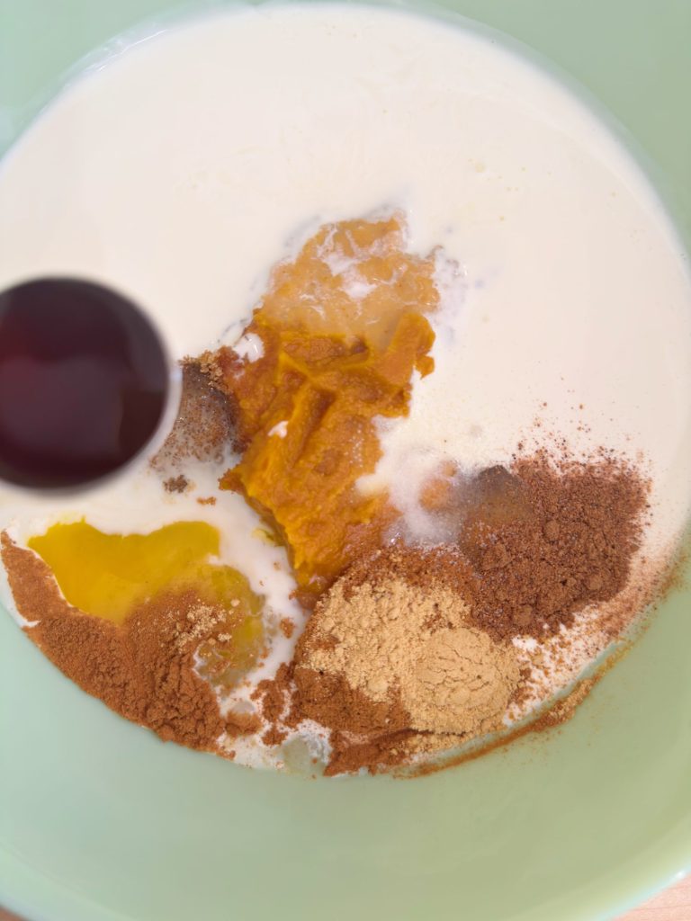 A mixing bowl containing cream, pumpkin puree, eggs, ground cinnamon, ginger, and a spoon with vanilla extract being added.