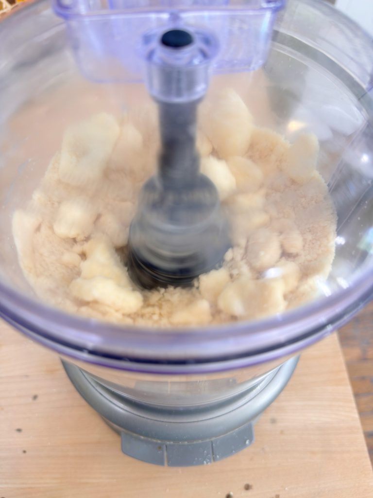 A food processor blending light-colored ingredients, possibly banana slices and flour, on a wooden surface.