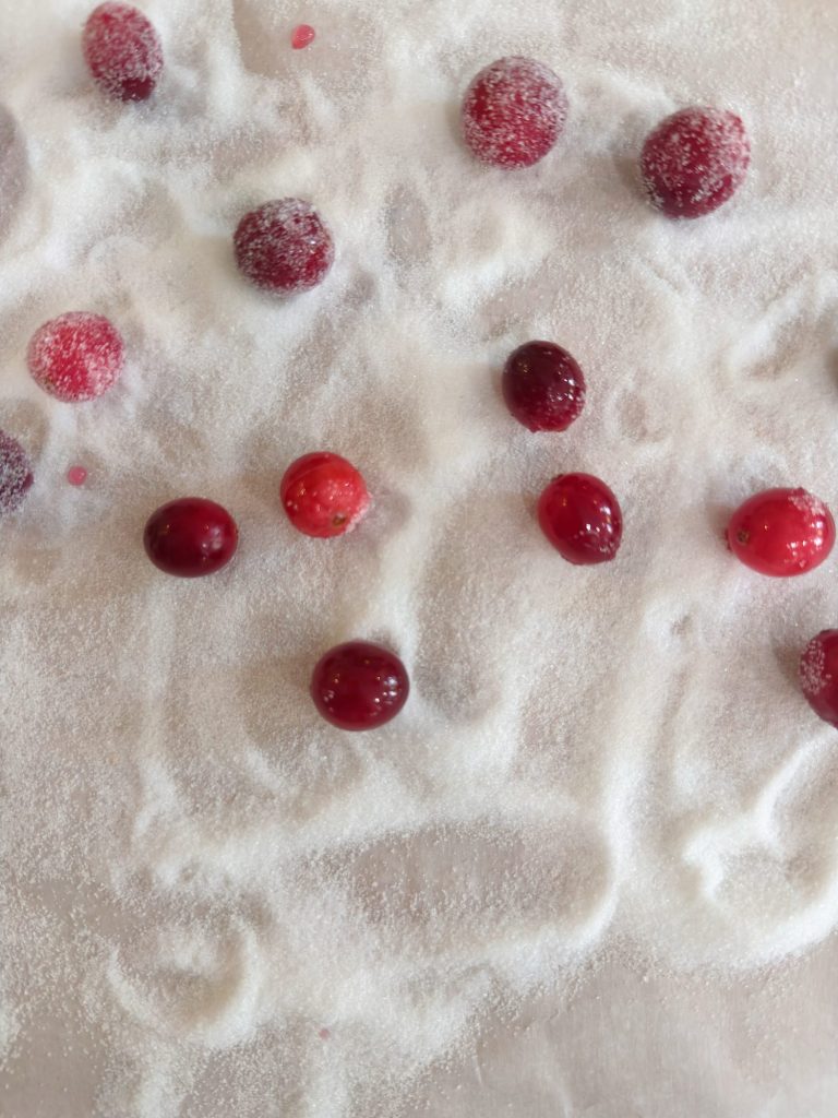 Red cranberries scattered on a white surface coated with granulated sugar, some berries partially covered by sugar.