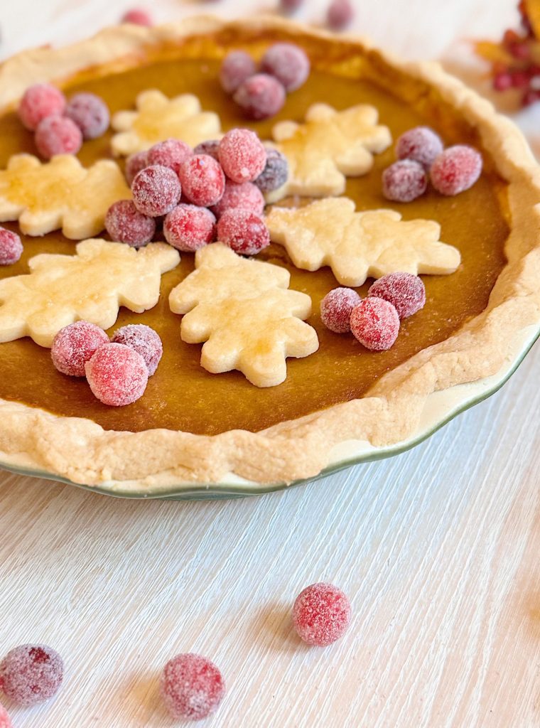 A pumpkin pie topped with sugared cranberries and leaf-shaped pie crust cutouts, displayed on a white surface.