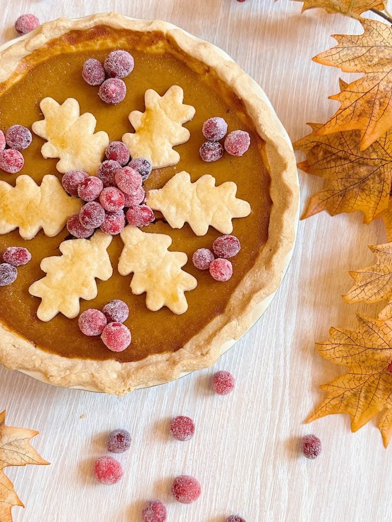 A pumpkin pie topped with sugared cranberries and leaf-shaped pie crust decorations, surrounded by autumn leaves and scattered cranberries on a light wood surface.