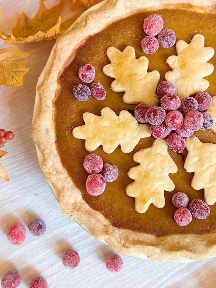 Pumpkin pie topped with leaf-shaped pastry cutouts and sugared cranberries, surrounded by autumn leaves and loose cranberries on a light surface.