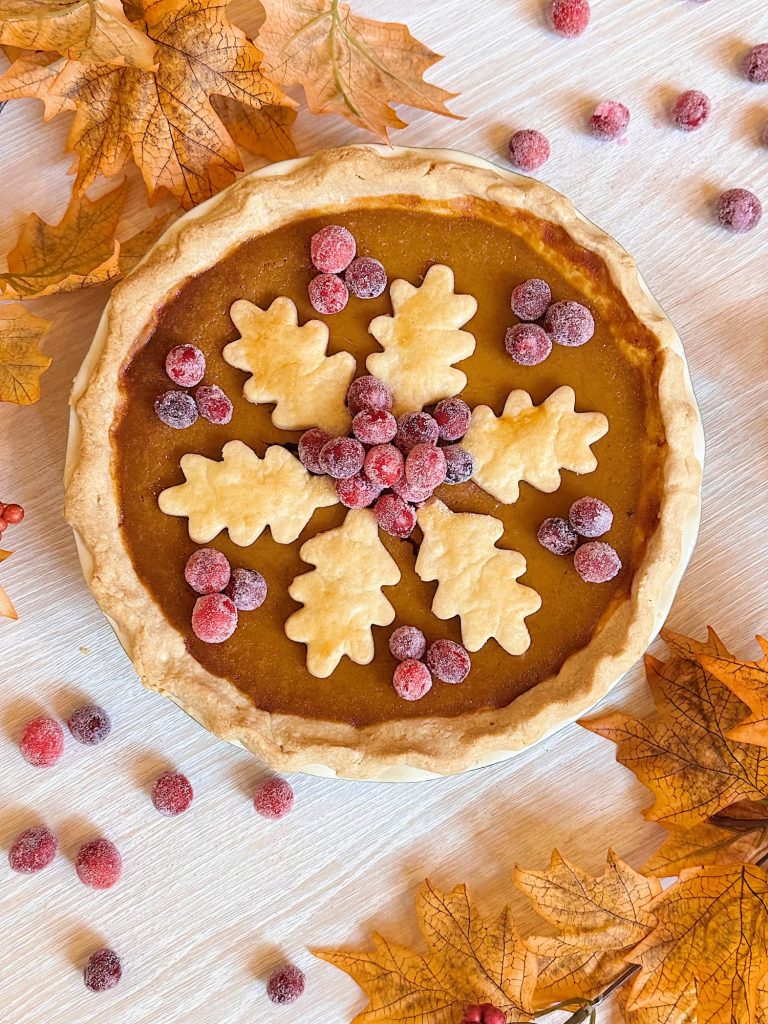 A pumpkin pie topped with leaf-shaped pastry cutouts and sugared cranberries, surrounded by scattered autumn leaves and cranberries on a light wooden surface.