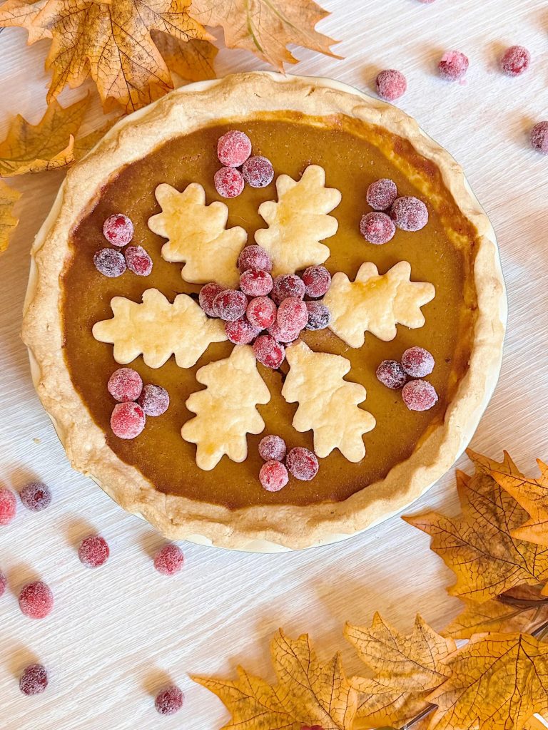 Pumpkin pie topped with leaf-shaped pie crust cutouts and sugared cranberries, surrounded by autumn leaves and cranberries on a light surface.