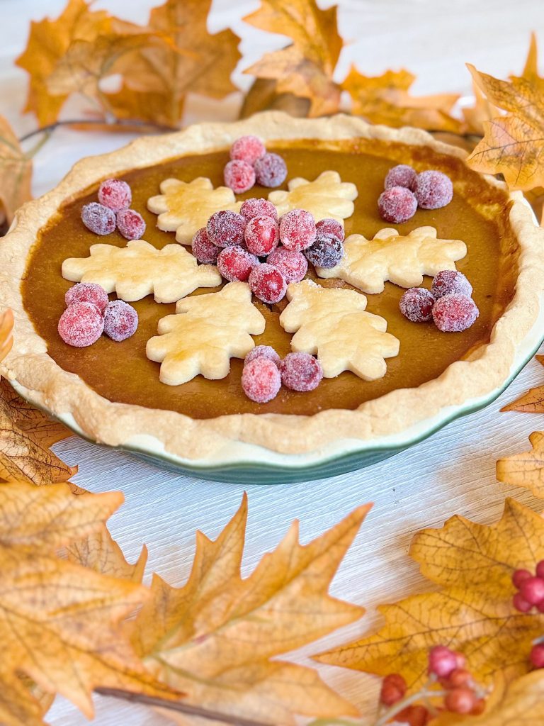 A pumpkin pie decorated with leaf-shaped pastry and sugared cranberries, surrounded by artificial autumn leaves on a white surface.