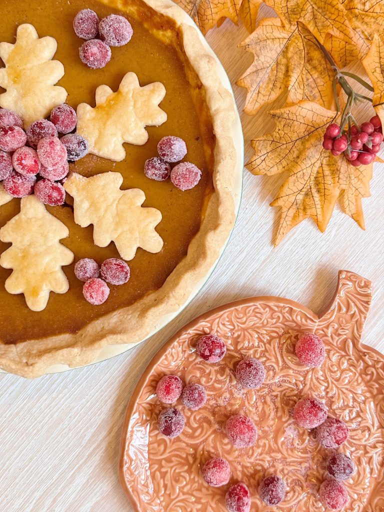 Pumpkin pie topped with leaf-shaped pastry and sugared cranberries, next to an orange leaf-shaped plate with more cranberries and artificial autumn leaves.