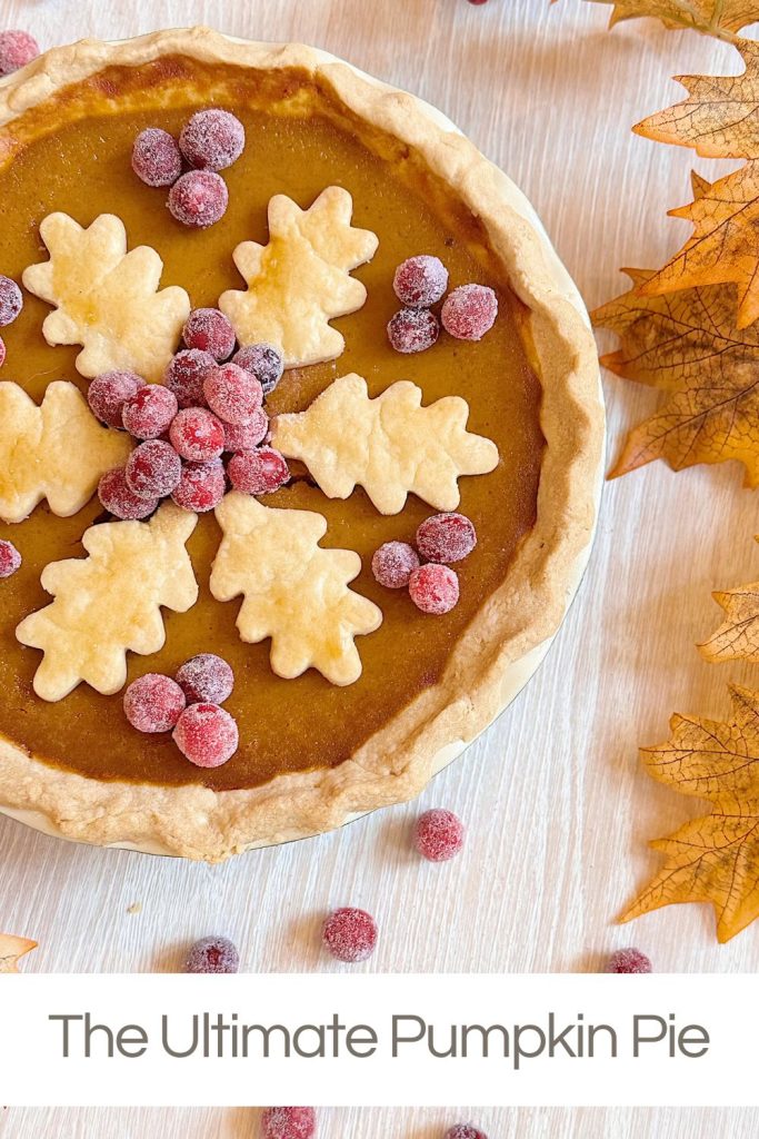 Pumpkin pie topped with leaf-shaped pastry and sugared cranberries, surrounded by autumn leaves on a light wooden surface. Text below reads: "The Ultimate Pumpkin Pie.