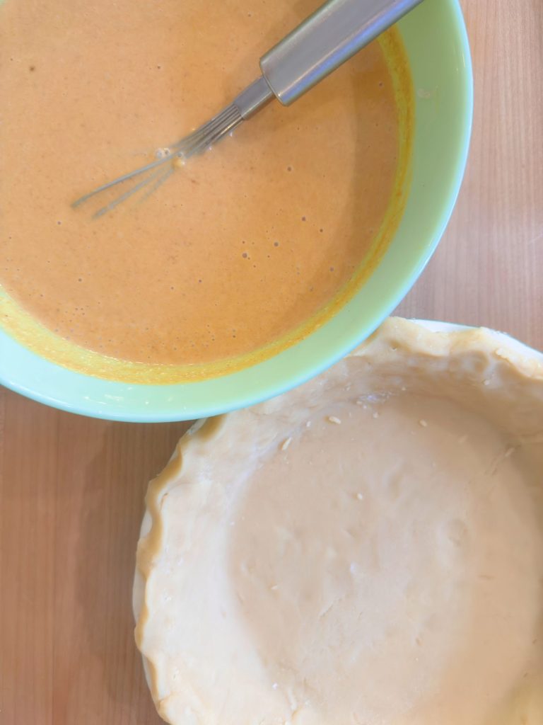 A bowl of pumpkin pie filling with a whisk beside an unbaked pie crust on a wooden surface.