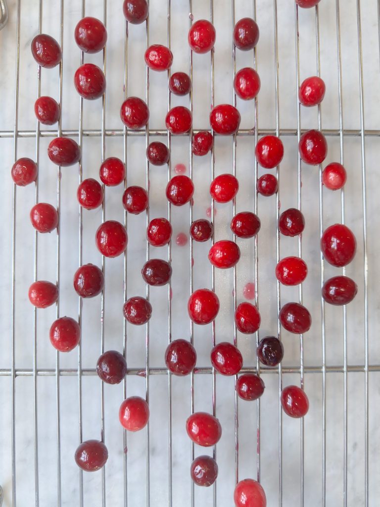 Glossy red and dark red cranberries are placed on a metal cooling rack, with a white surface underneath.
